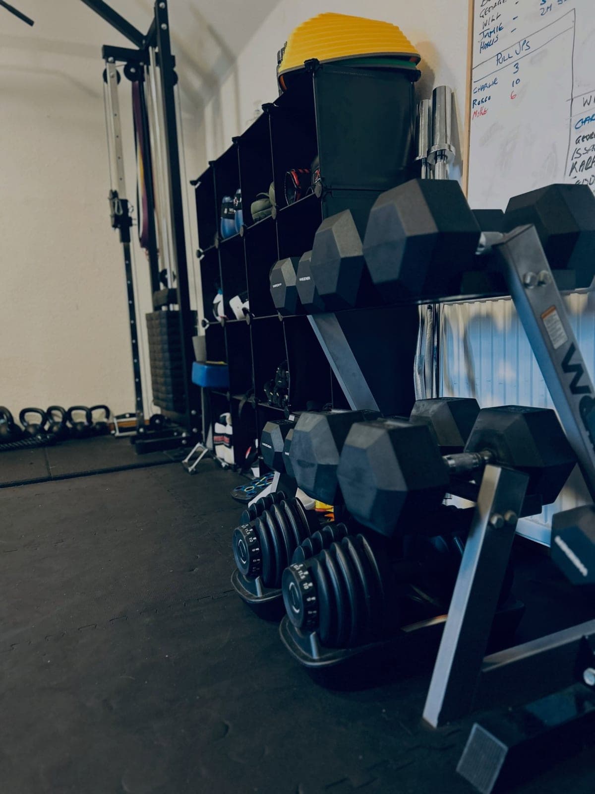 The dumbbell rack and kettlebells, beside the whiteboard with the week's names and numbers on it