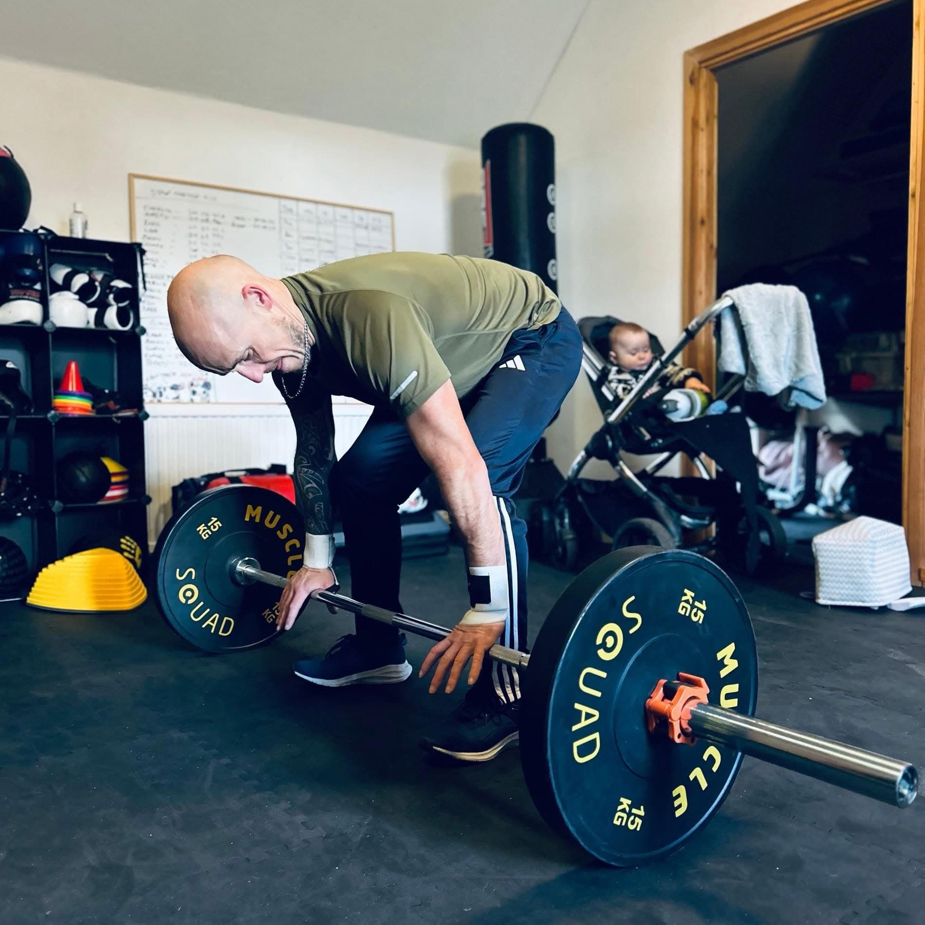 Ross setting up a deadlift in his gym in Elgin, the session whiteboard behind him and his youngest watching from a pushchair