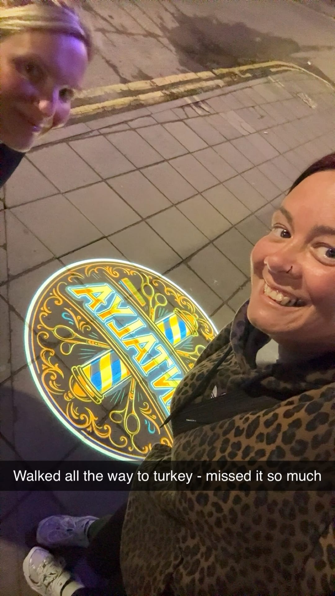 Sarah and a friend laughing beside a brightly lit barber's sign at night